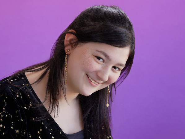 A person with long dark hair is smiling while wearing a black top with star patterns and long star-shaped earrings. The background is a solid purple color.