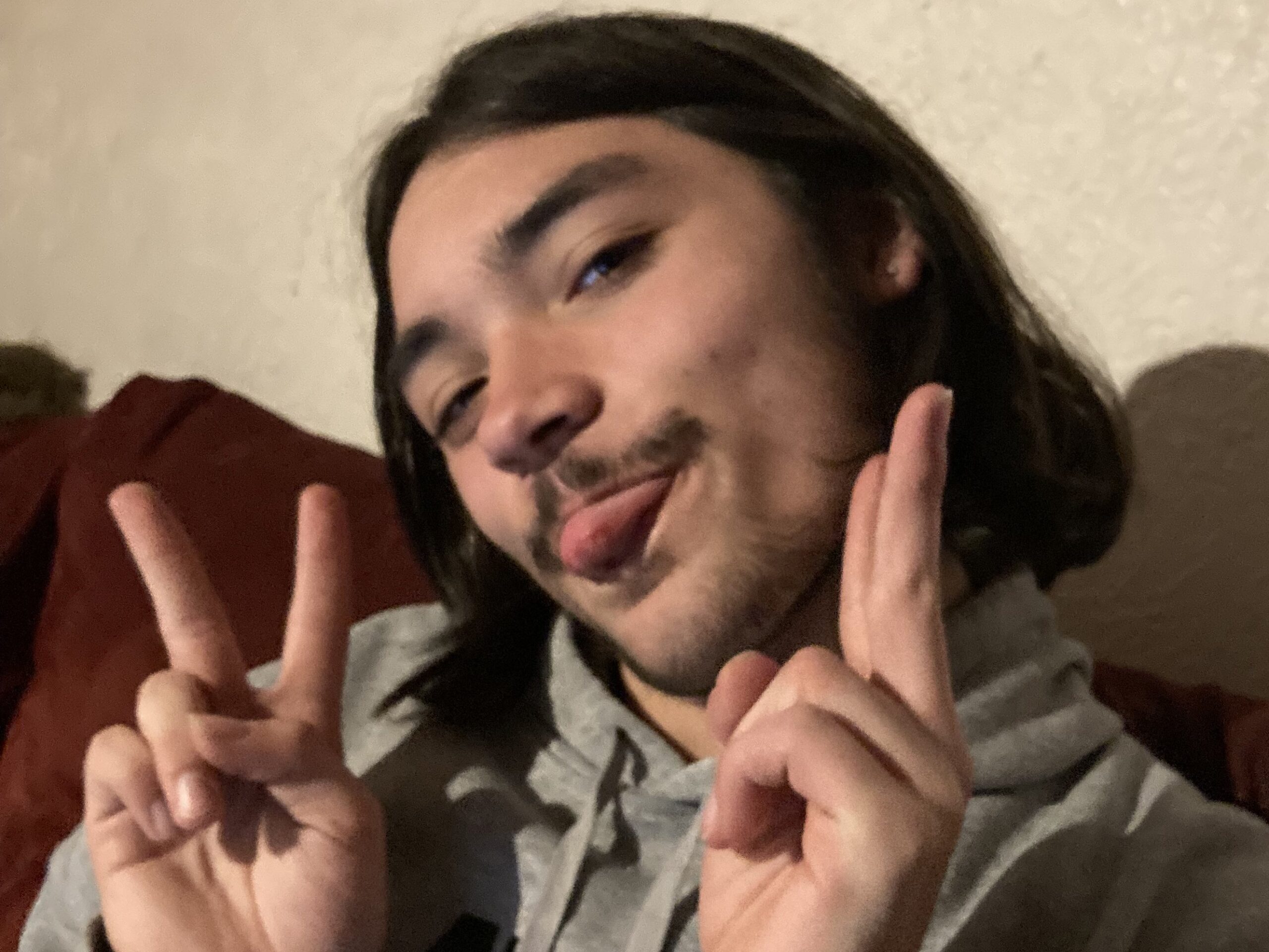 A person with shoulder-length brown hair poses playfully, sticking out their tongue and holding up two peace signs with their fingers. They are wearing a gray hoodie and are in a room with a beige wall.