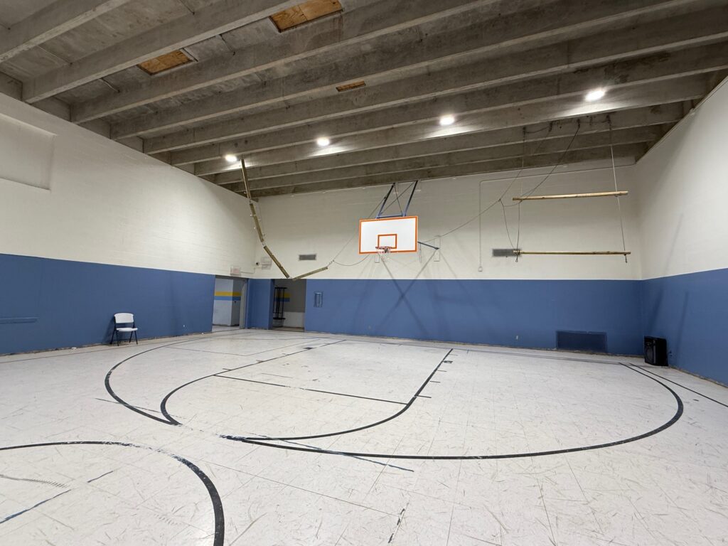 A mostly empty indoor gymnasium with a basketball hoop mounted on the far wall, faded lines on the floor, blue and white walls, exposed ceiling beams, and a single white chair by the wall.