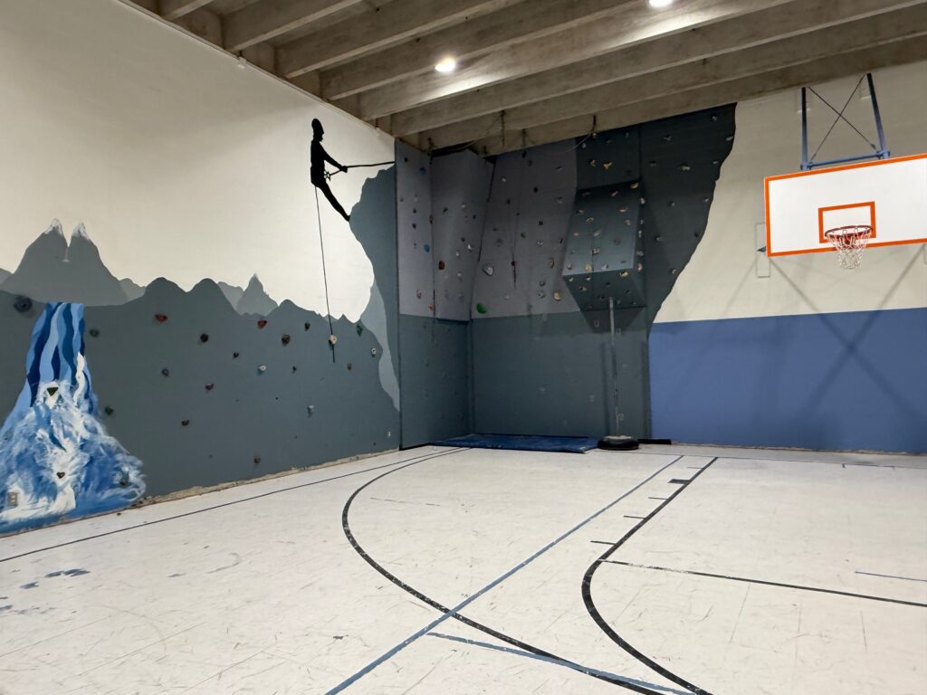 Indoor gym with a rock climbing wall in the corner, a mural of a climber and a waterfall on the wall, and a basketball hoop mounted above the court. The floor markings indicate a basketball court.