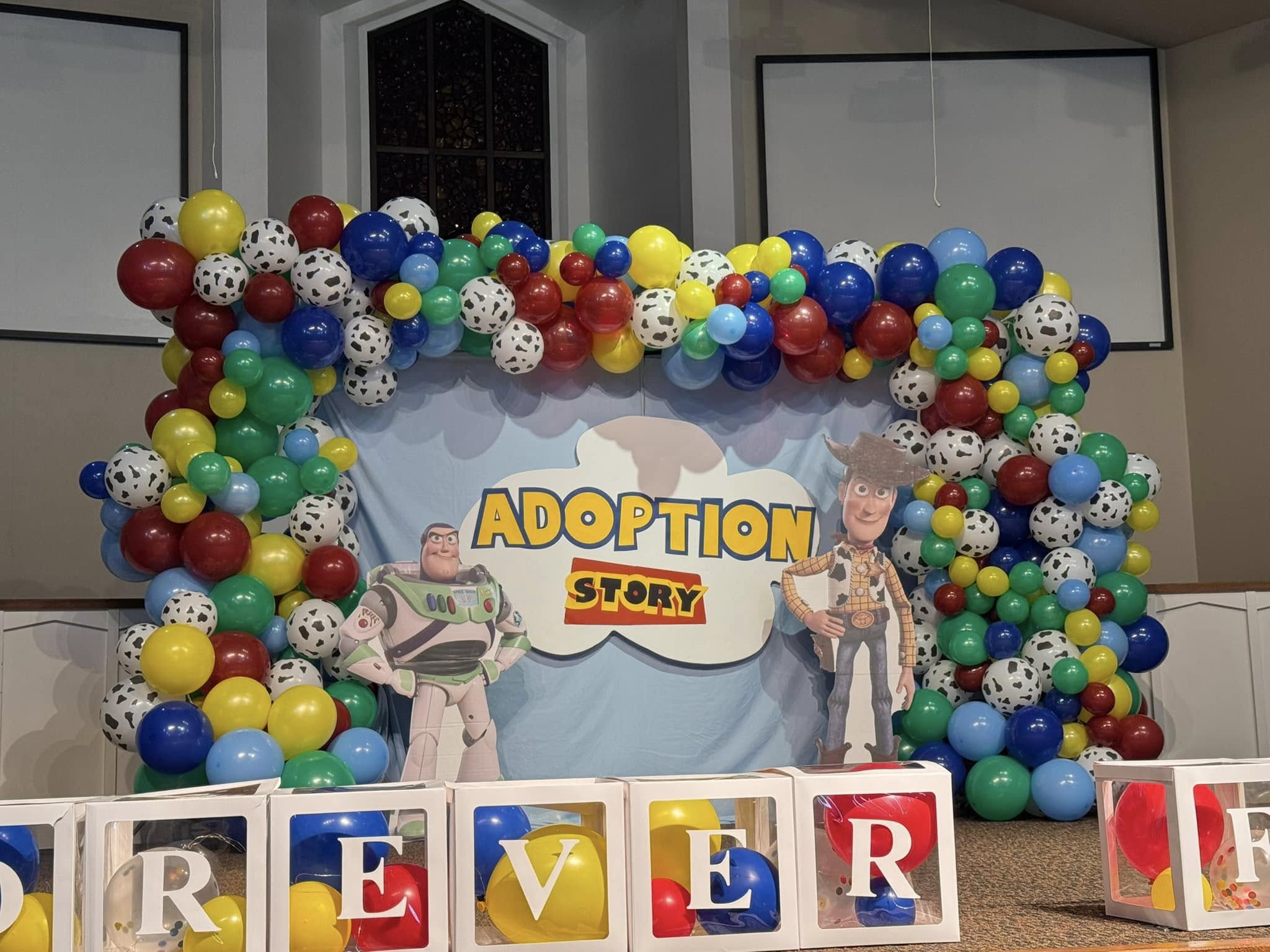 A colorful balloon arch with red, yellow, blue, green, and black-and-white spotted balloons frames a backdrop with "Adoption Story" text. Cardboard cutouts of two animated characters are in front, alongside large blocks spelling "FOREVER.