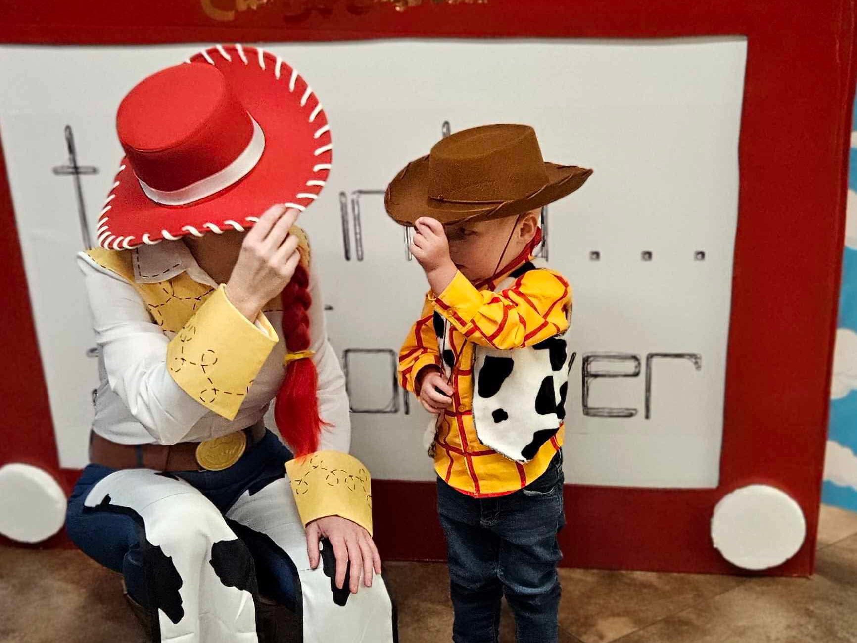 Two people in cowboy-themed costumes are tipping their hats. One is dressed in a red and white outfit with a red hat and braid, and the other, a child, is in a yellow shirt with cow print and a brown hat. A large toy sketch board is in the background.