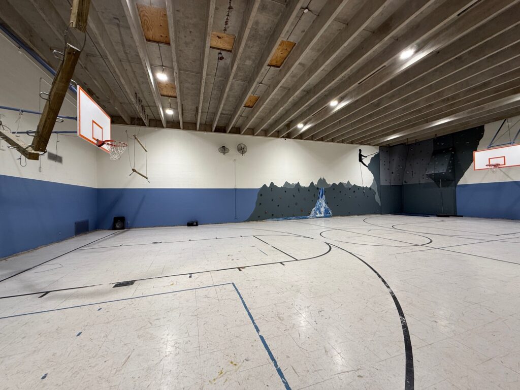 Indoor gymnasium with a white and blue floor, two basketball hoops, concrete ceiling, and a rock climbing wall on the far side. The gym is empty and well-lit with overhead lights.