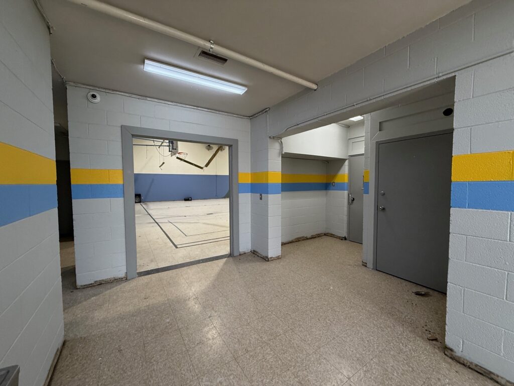 A hallway with beige tile floors and gray doors leads into an empty gymnasium. The cinder block walls are painted white with blue and yellow stripes. Overhead fluorescent lighting illuminates the space.