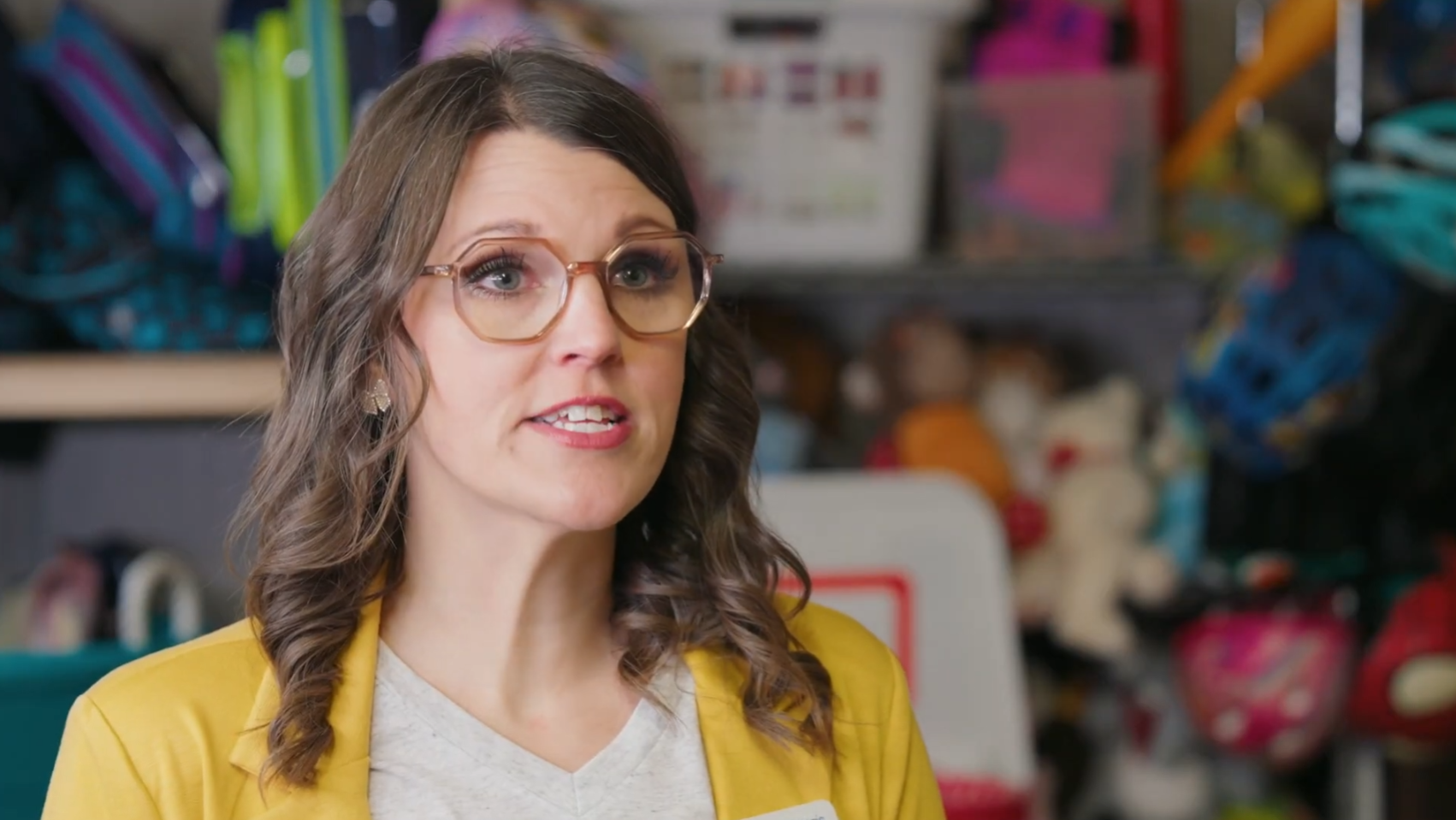 A woman with brown curly hair and glasses is speaking. She wears a yellow blazer over a gray shirt. Behind her are assorted colorful items on shelves, including a white bin and various bags and toys.