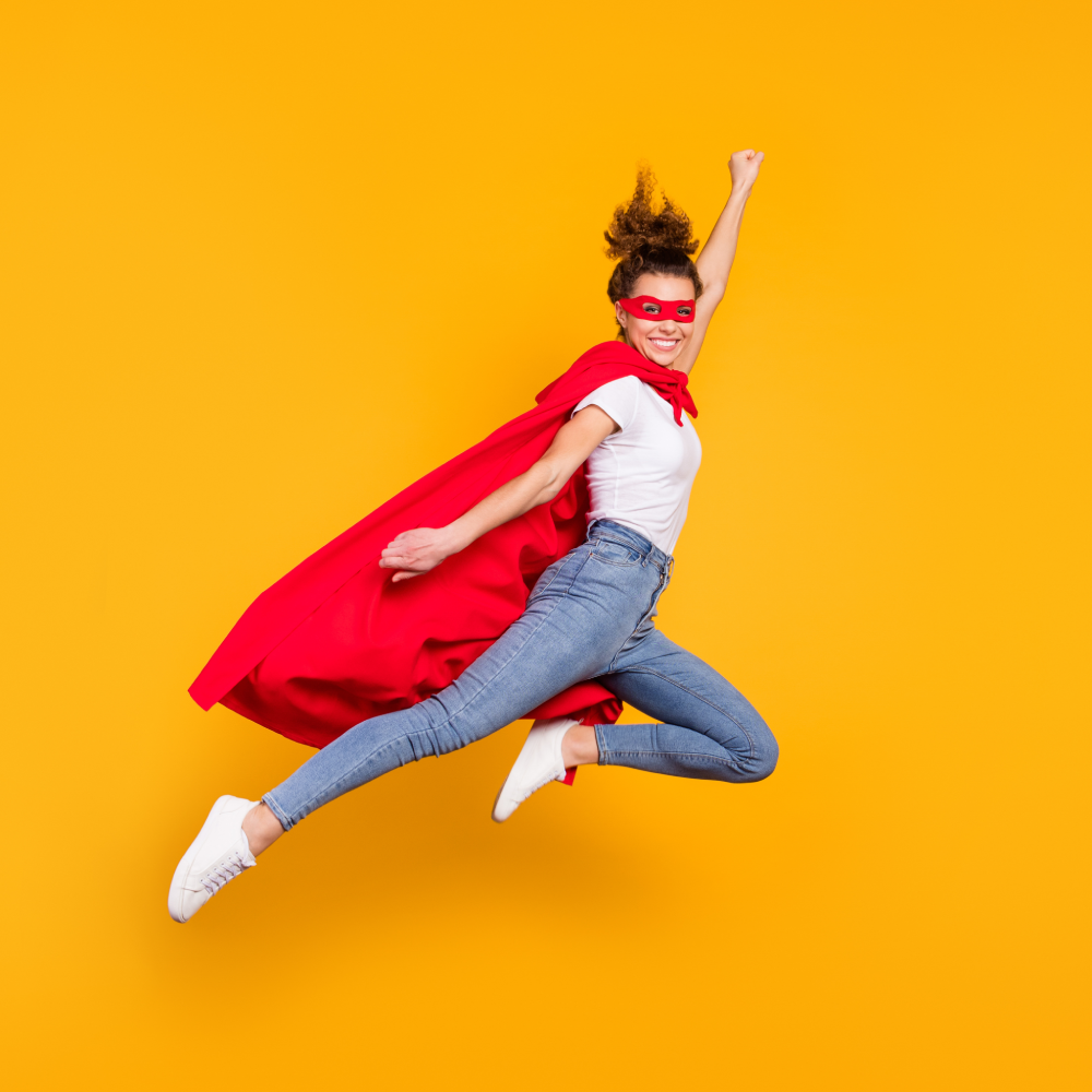 A woman wearing a red cape and red mask, white t-shirt, blue jeans, and white sneakers jumps in the air against a solid yellow background, posing with one arm raised as if flying.
