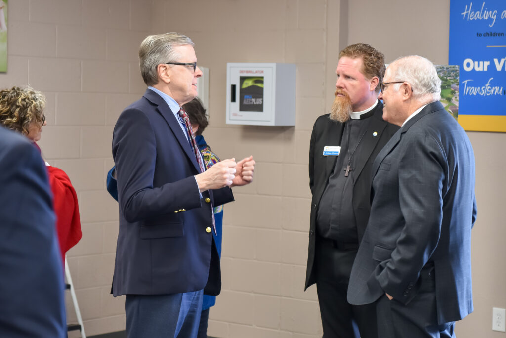Three men in business attire, including one wearing a clerical collar, are standing indoors having a conversation. One man gestures with his hands while the others listen. A wall-mounted defibrillator and signage are visible in the background.