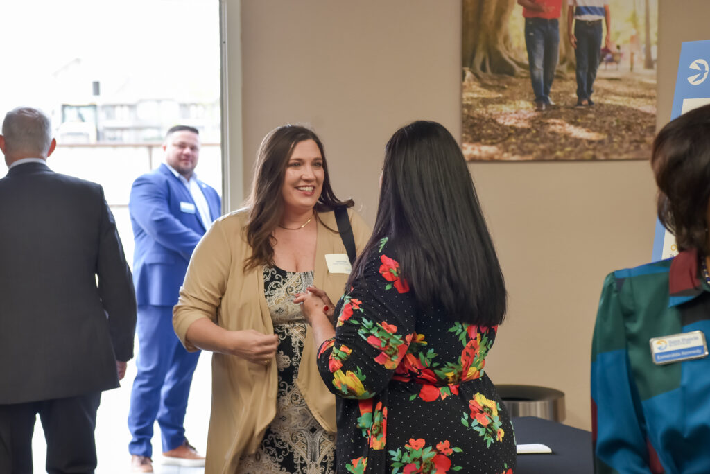 Two women stand talking at an indoor event, with other people visible in the background. One woman wears a floral dress and the other a tan cardigan. A poster and framed photo are seen on the wall behind them.