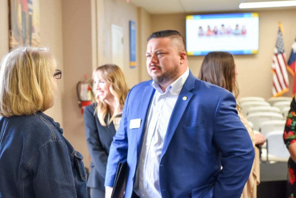A man in a blue suit jacket speaks with a woman wearing glasses and a denim jacket. Other people stand and talk in the background. An American flag and chairs are visible, along with a screen displaying images.