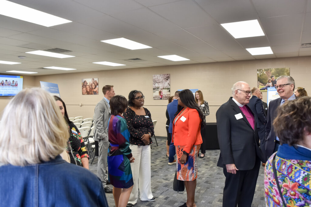 A group of people in business or formal attire stand and converse in a brightly lit room with chairs arranged in rows and photos on the walls.