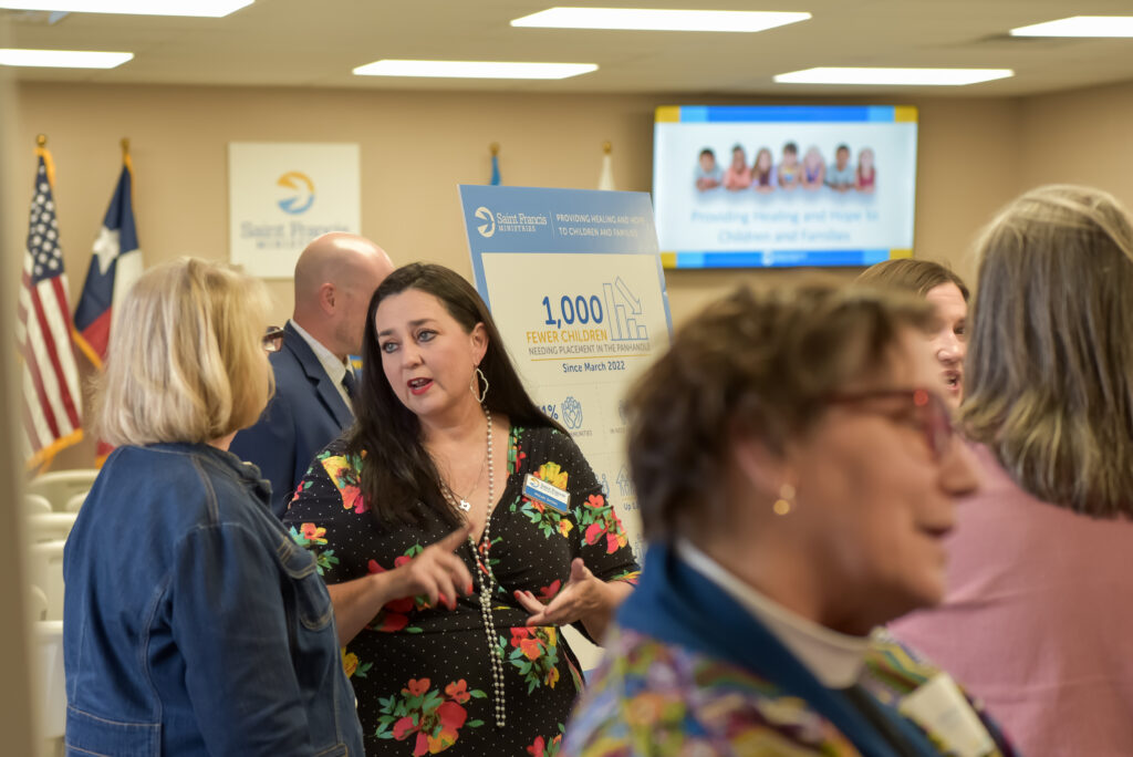 A group of adults converse in a meeting room. A woman in a floral dress stands near a poster displaying statistics. American and Texas flags are in the background along with signs and a screen showing a family photo.