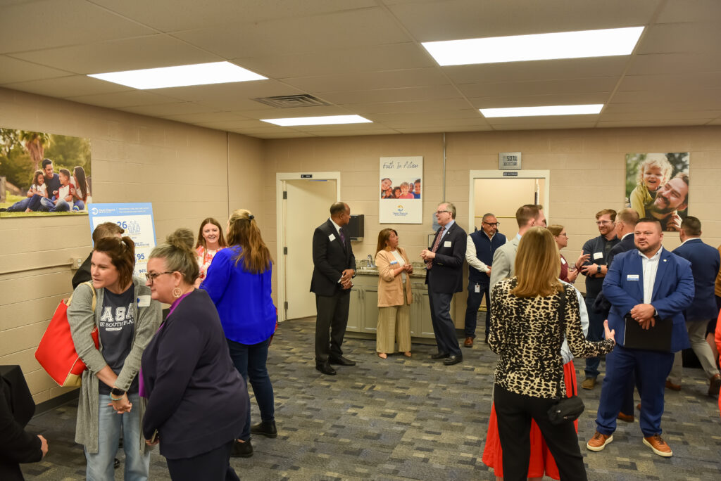 A group of adults in business attire converse in small groups in a carpeted hallway with beige walls decorated with family photos and informational posters. Some people hold bags or paper folders.