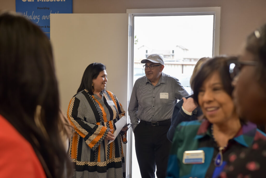 A group of adults gathers indoors near an open door. A woman in a patterned dress is talking to a man in a cap and glasses. Others stand in the foreground, some wearing name tags.