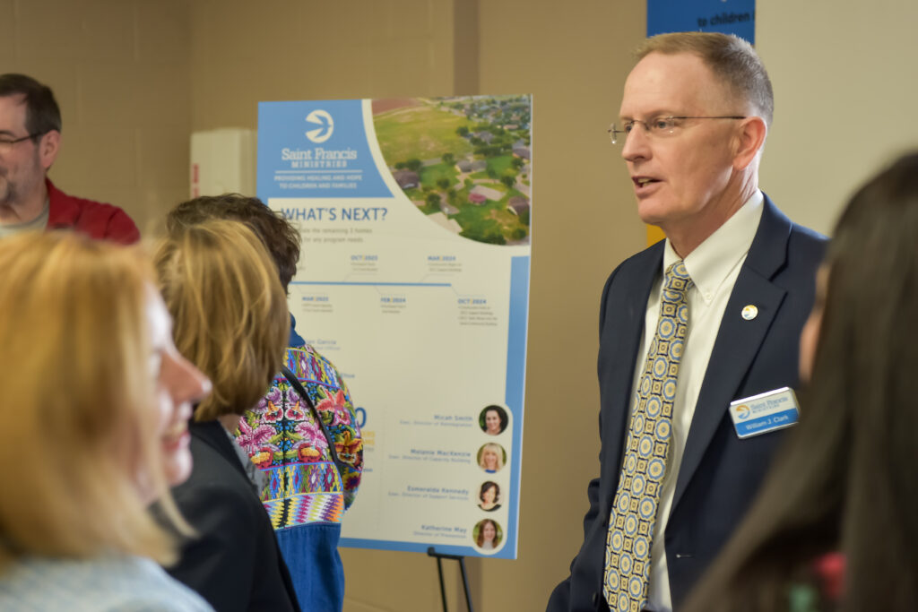 A man in a suit and tie, wearing a name badge, stands and talks with a group of people near a display board labeled "Saint Francis" and "What's Next?" The background includes other attendees and informational posters.