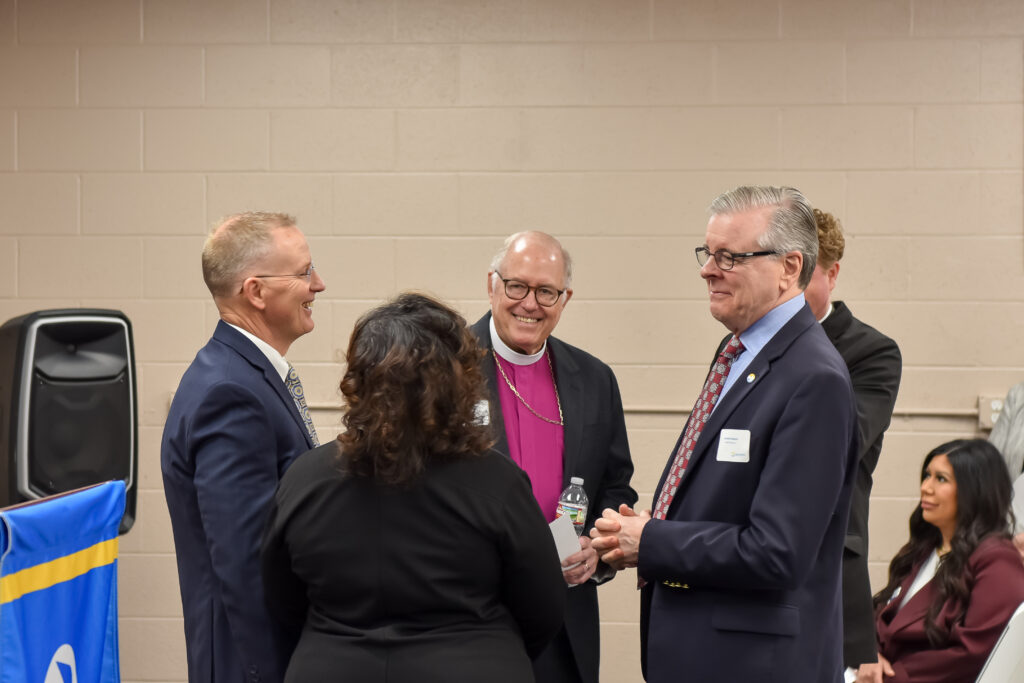 Four adults in formal attire stand in conversation in a room with tan brick walls. One man in a purple clerical shirt and cross necklace smiles at another man in glasses and a suit. Two people have their backs to the camera.