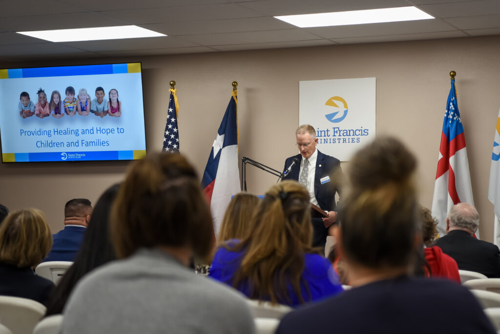 A man stands at a podium addressing a seated audience in a conference room. Behind him are three flags, a Saint Francis Ministries sign, and a screen displaying "Providing Healing and Hope to Children and Families.