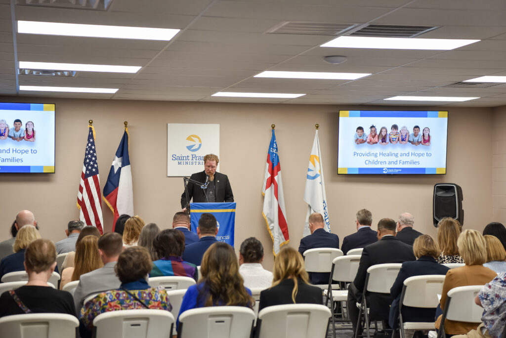 A man speaks at a podium in a conference room with seated attendees. Behind him are flags and two screens displaying the text “Providing Healing and Hope to Children and Families.”.