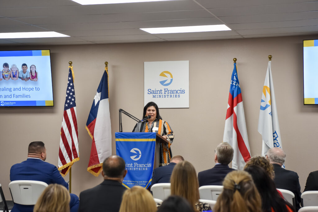 A woman speaks at a podium with a Saint Francis Ministries banner in front of an audience. Behind her are four flags, and a screen displaying a photo of three children and text about healing and hope for children and families.