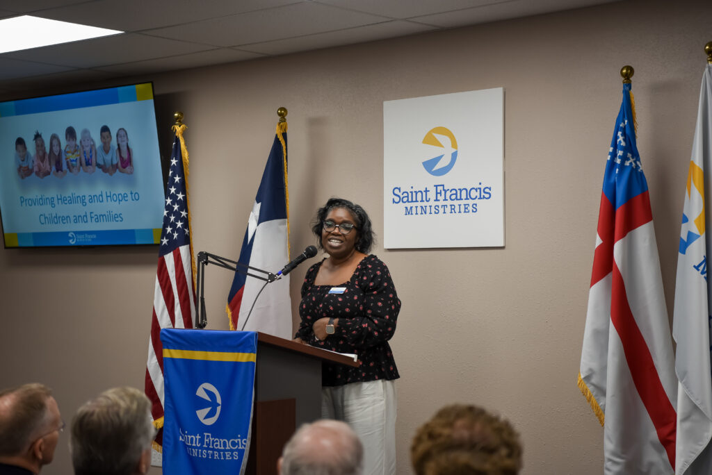 A woman stands at a podium giving a speech in a room with Saint Francis Ministries signs, American and state flags, and an audience. A screen displays text about providing healing and hope to children and families.
