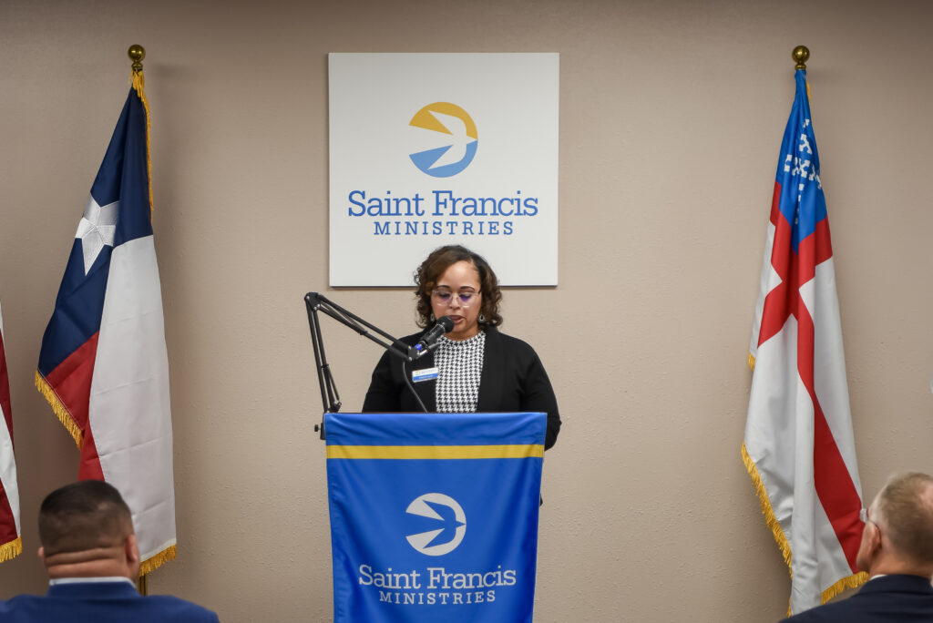 A woman stands at a podium with a microphone, speaking in front of a “Saint Francis Ministries” sign. Two flags are on either side of her, and several people are seated facing her.