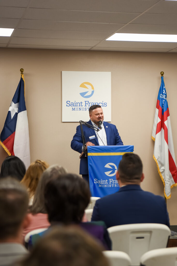 A man in a suit stands at a podium with a Saint Francis Ministries banner, speaking to an audience. Two flags are displayed behind him, and a Saint Francis Ministries sign is on the wall.