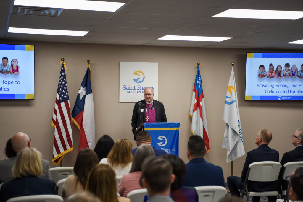 A man in religious attire speaks at a podium with a Saint Francis Ministries logo, flanked by U.S. and Texas flags, to an audience in a conference room. Two screens display a message about hope for children and families.