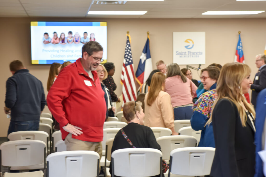 Several people are standing and talking in a meeting room filled with white chairs. Three flags and a presentation screen with the Saint Francis Ministries logo are visible in the background.