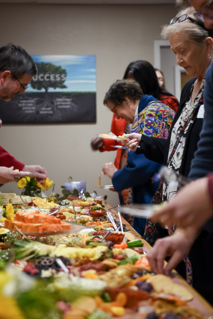 A group of people stands around a table filled with assorted foods, serving themselves at a buffet-style gathering in an indoor setting. A SUCCESS poster is visible on the wall in the background.