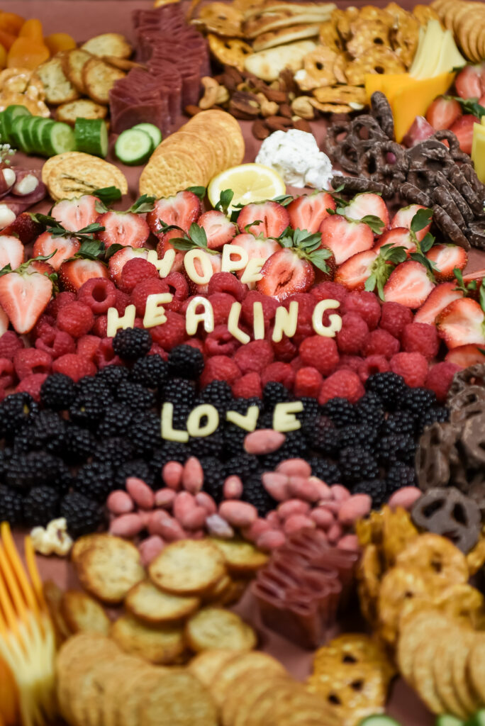 A charcuterie board arranged with crackers, cheese, chocolate, strawberries, raspberries, and blackberries. Pieces of cheese spell out "JOY," "HEALING," and "LOVE" among the fruit in the center.