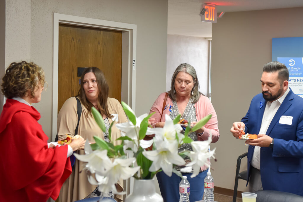 Four adults stand indoors, conversing and holding plates of food. A white vase with lilies is on the table in the foreground. Two women wear cardigans, while a man in a suit and a woman in red are present. Bottled water is visible.