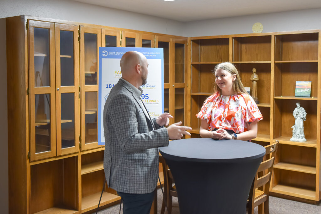 A man and a woman stand talking at a round, tall table in front of wooden bookshelves with glass doors. The man gestures with his hands while the woman listens and smiles. A poster is visible in the background.