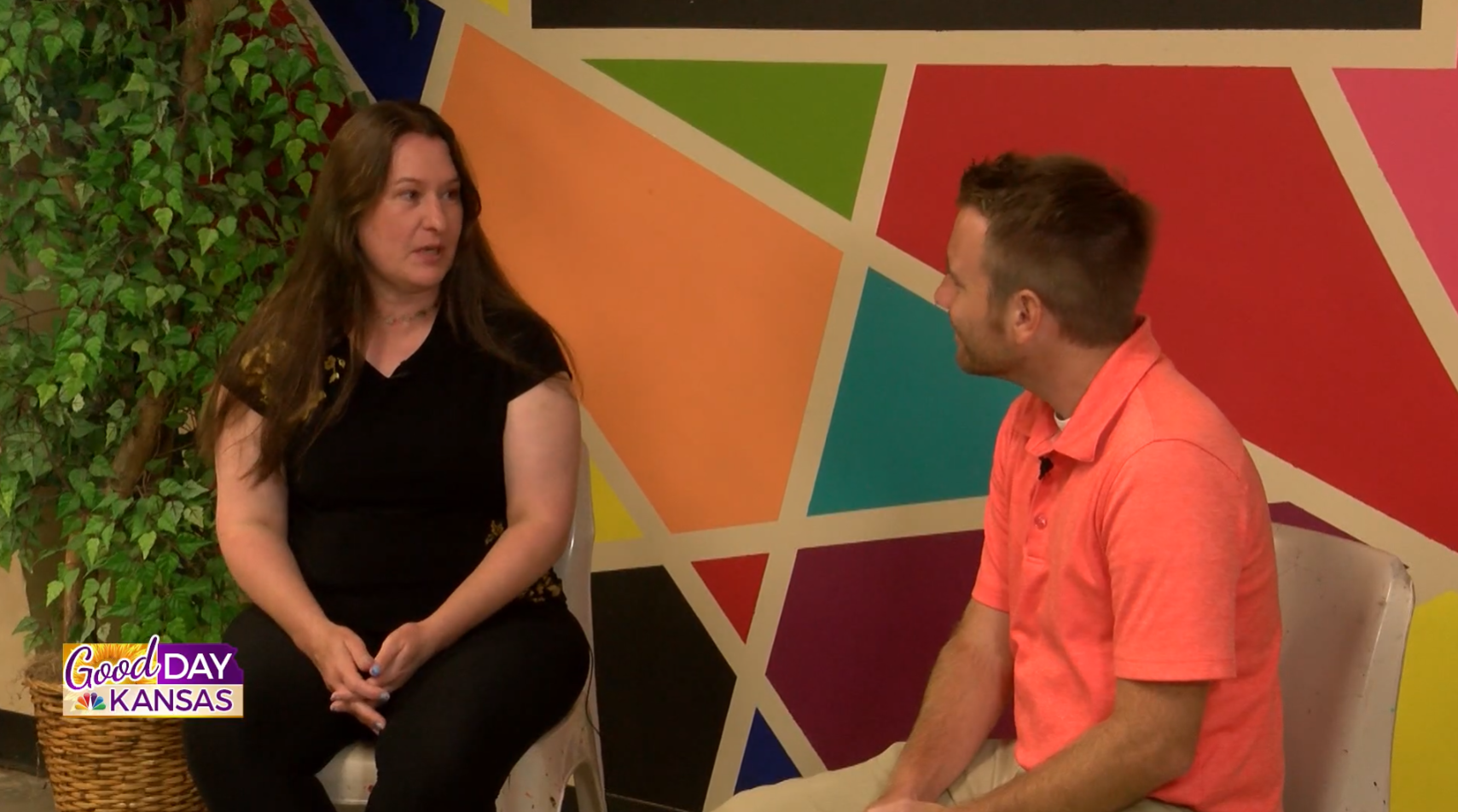 A woman in black and a man in a coral shirt sit and talk in front of a colorful geometric wall. A “Good Day Kansas” graphic is visible in the lower left corner.