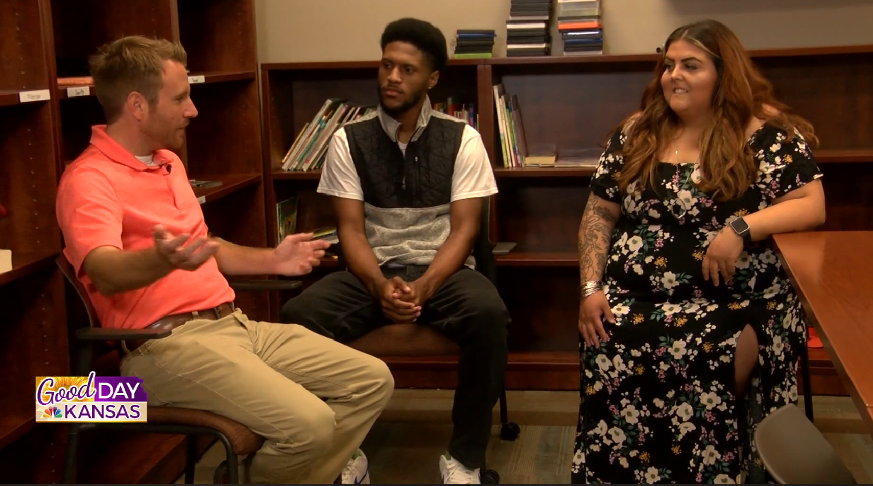 Three people sit and talk in a library or office setting. The man on the left gestures with his hands, while the man in the middle and the woman on the right listen. A "Good Day Kansas" logo is visible in the lower left corner.