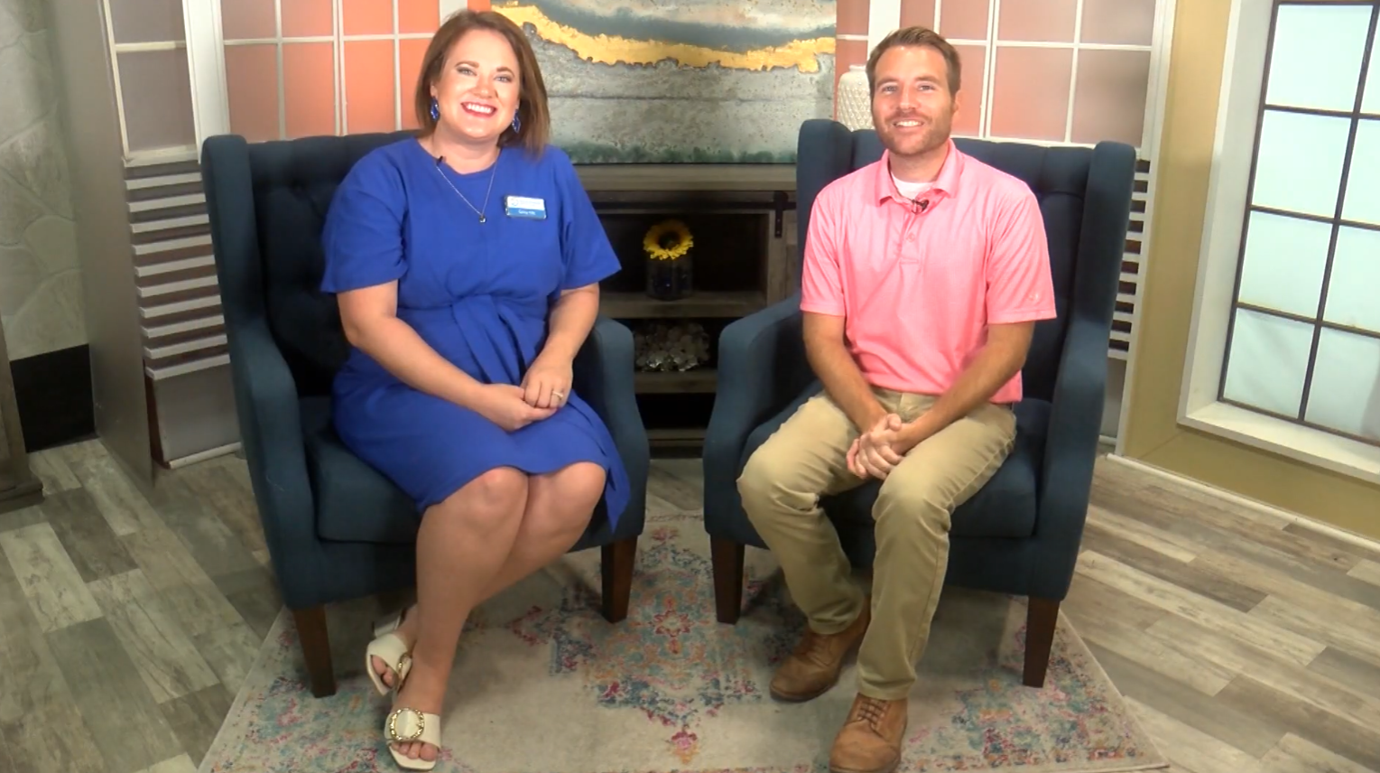 A woman in a blue dress and a man in a pink polo shirt sit on dark blue chairs in a brightly lit studio with a modern decor and a decorative rug on the floor. Both are smiling and looking at the camera.