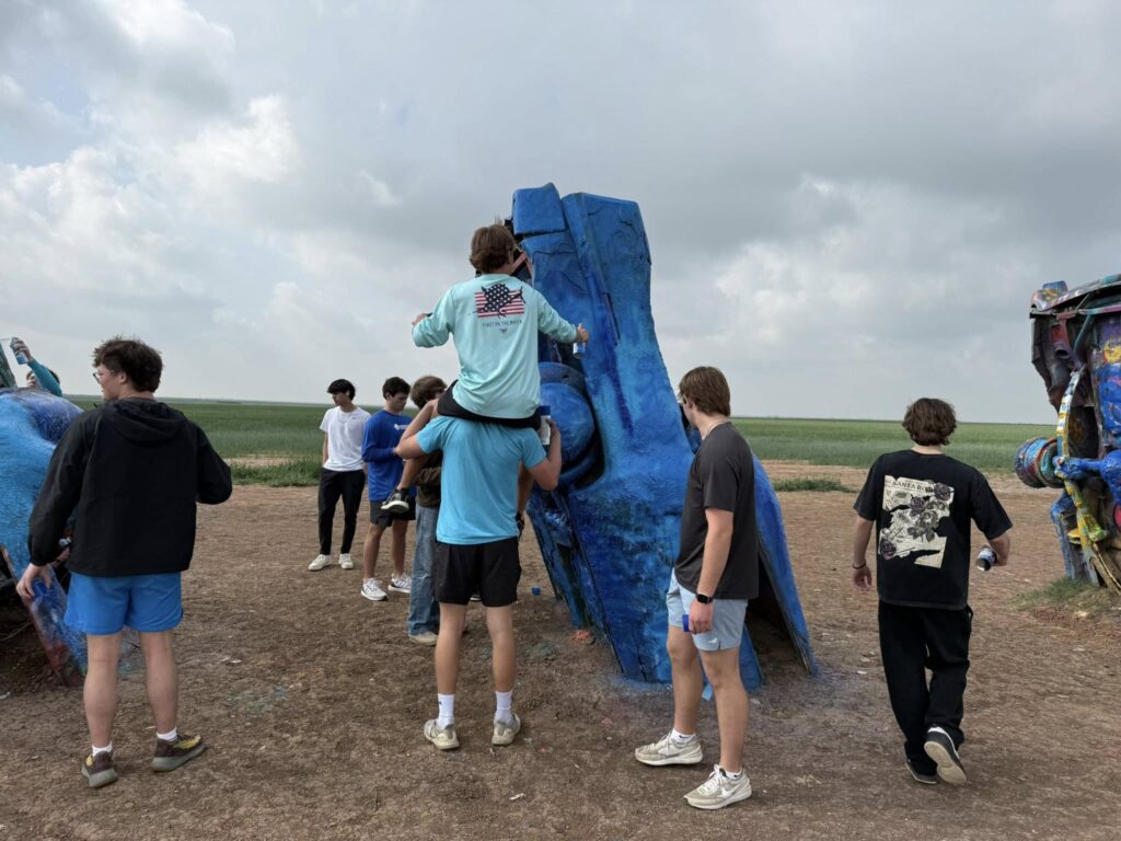 A group of young people stand around and interact with a large, blue-painted car partially buried upright in the ground in an open outdoor area under a cloudy sky.