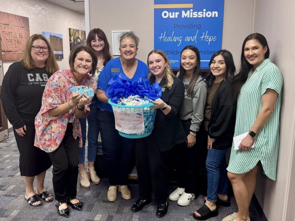 Eight women stand together indoors, smiling at the camera. One holds a large blue and white gift basket. Behind them is a sign that says “Our Mission: Providing Healing and Hope for Children and Families.”.