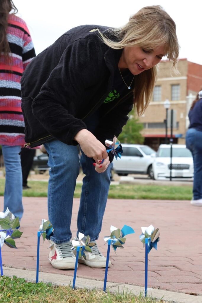 A woman in a black jacket and jeans bends down to place a blue and silver pinwheel in the ground alongside several others on a sidewalk, with parked cars and people visible in the background.