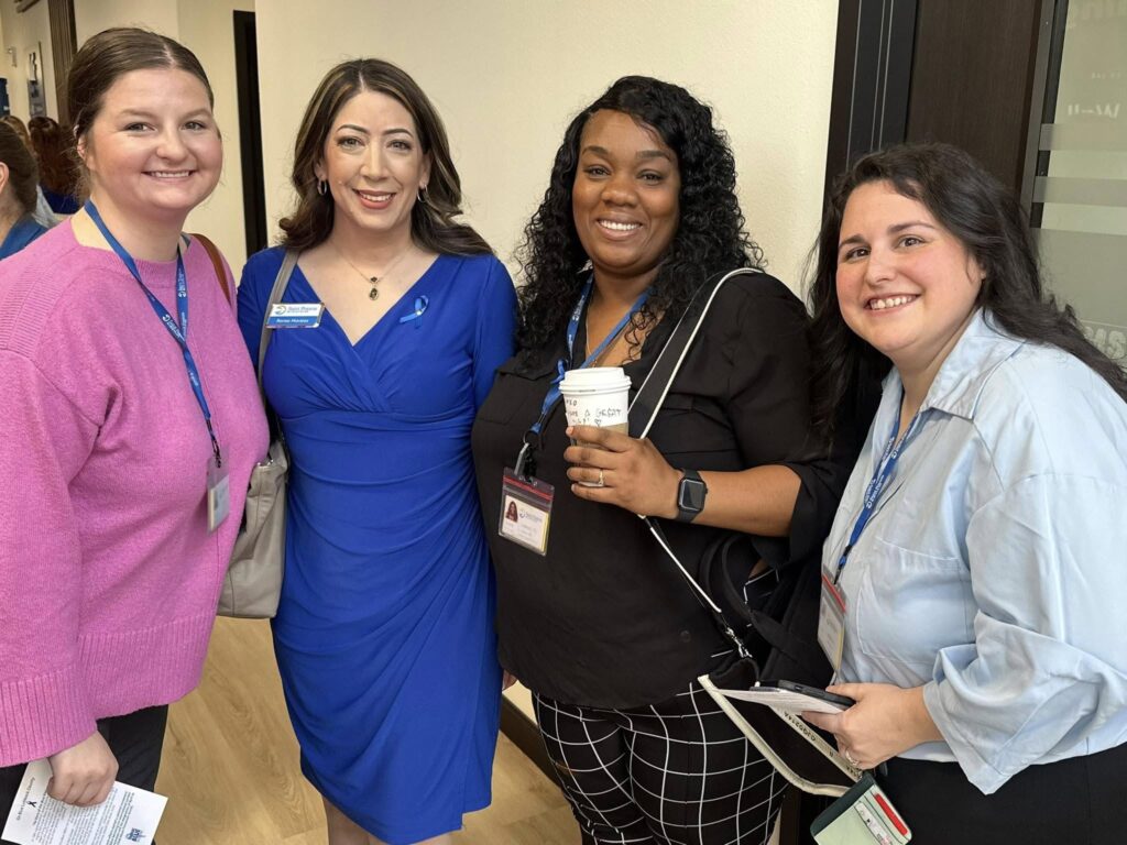 Four women stand together indoors, smiling at the camera. They wear business or business-casual attire and display conference name badges. One holds a coffee cup, while others hold papers or tablets.