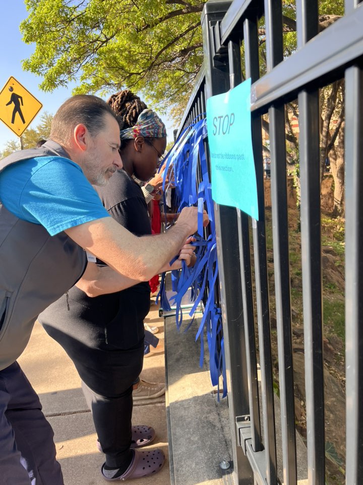 Two people tie blue ribbons to a black metal fence next to a sidewalk. A sign on the fence says "STOP" with instructions, and a pedestrian crossing sign is visible in the background near trees.