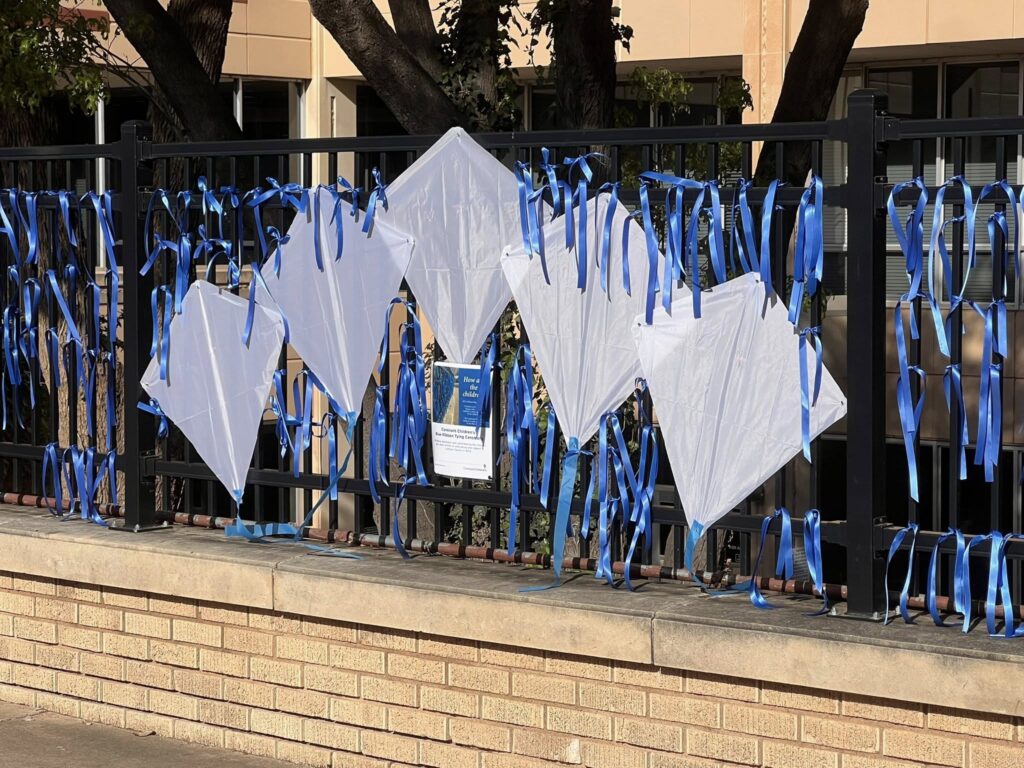Four white kites and several blue ribbons are attached to a black metal fence in front of a beige building. A small informational sign is posted on the fence among the kites and ribbons.