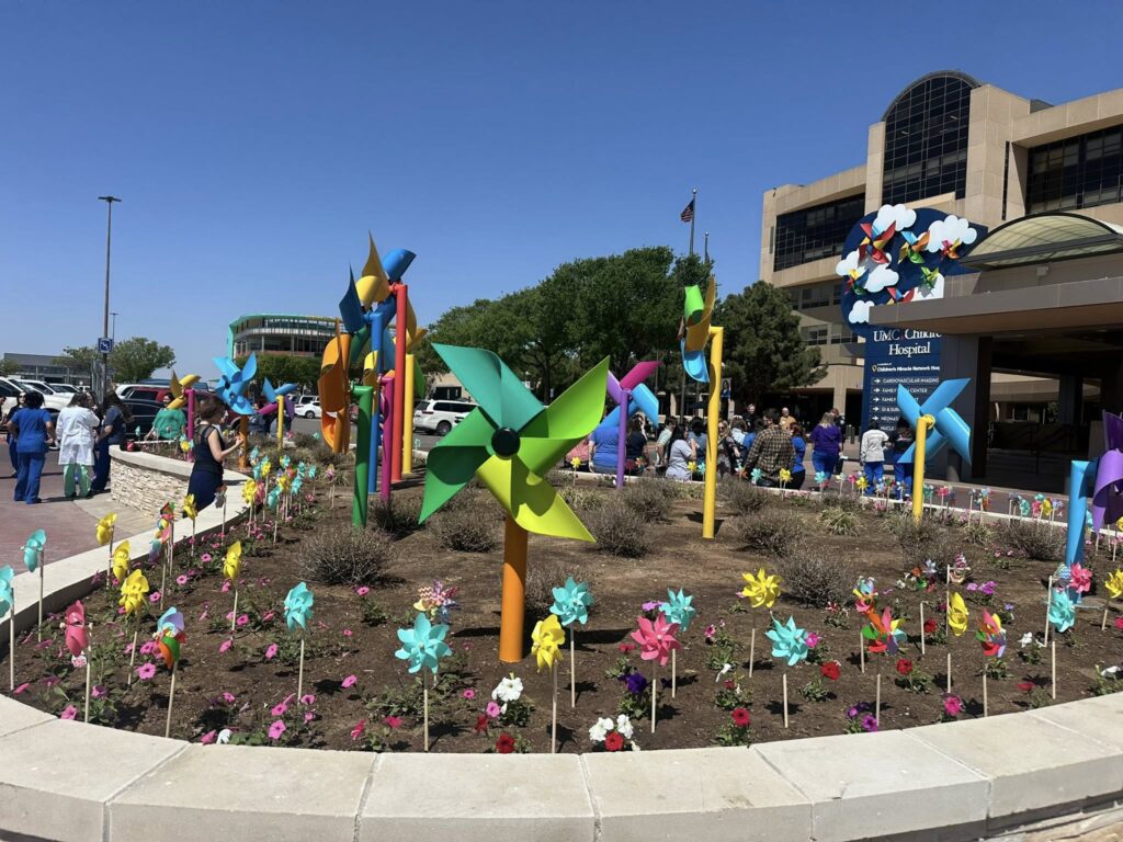 Large colorful pinwheels and small flower pinwheels are displayed in a circular garden bed outside a hospital building. People are gathered nearby, some wearing scrubs, under a clear blue sky.