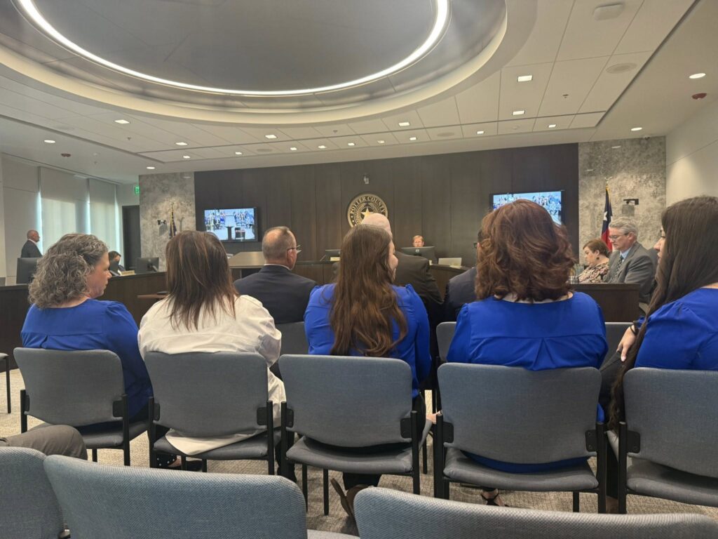 People sit in gray chairs inside a modern courtroom, facing a panel of judges. Several screens display virtual participants. Most of the women in the foreground wear blue tops. An American flag is visible to the right.