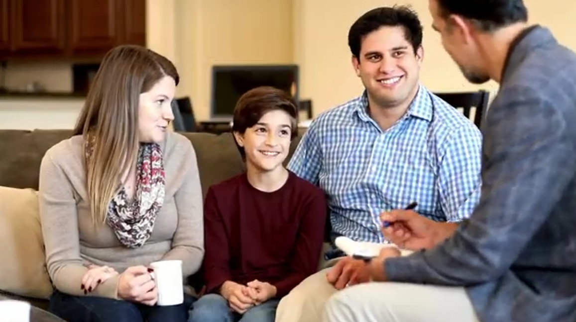 A woman, a boy, and a man sit on a couch facing another man who is talking to them and holding a pen and clipboard. They appear to be having a friendly discussion in a living room.
