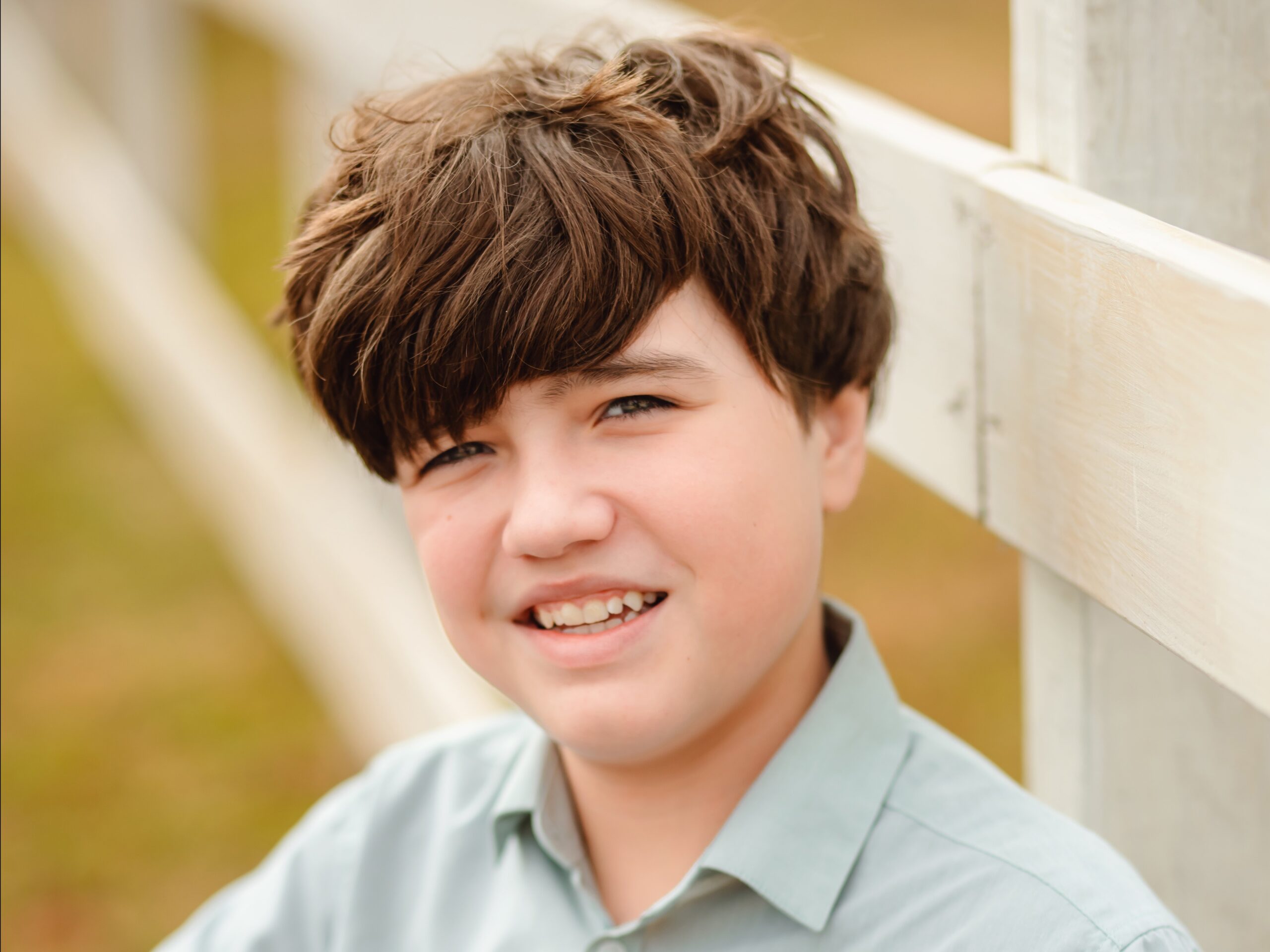 A boy with medium-length brown hair and a light green collared shirt smiles while standing in front of a white wooden fence outdoors. The background is blurred.