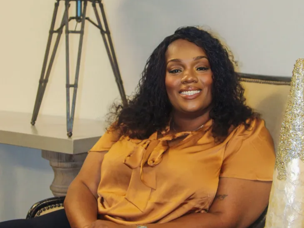 A woman with curly hair, wearing a mustard yellow blouse, is sitting on a chair and smiling at the camera. There is a small table with a decorative object behind her.